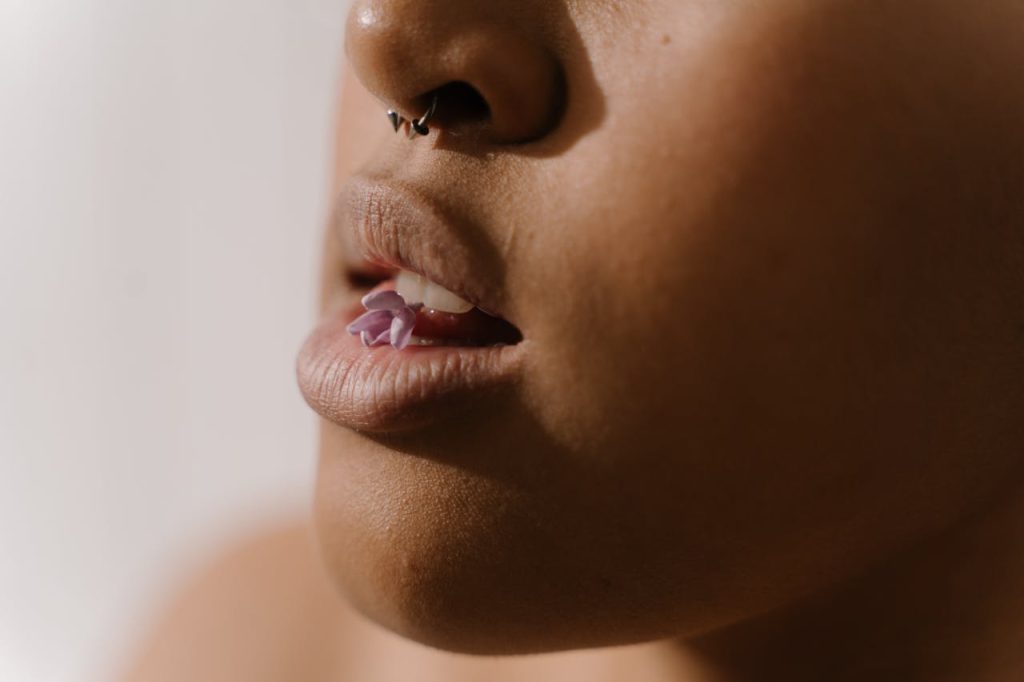 Artistic close-up of lips with a nose ring and a purple flower held between the teeth.